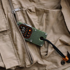 Apple Watch Ultra in an olive green miniPhone Ultra case resting on a tan vest beside a zipper.